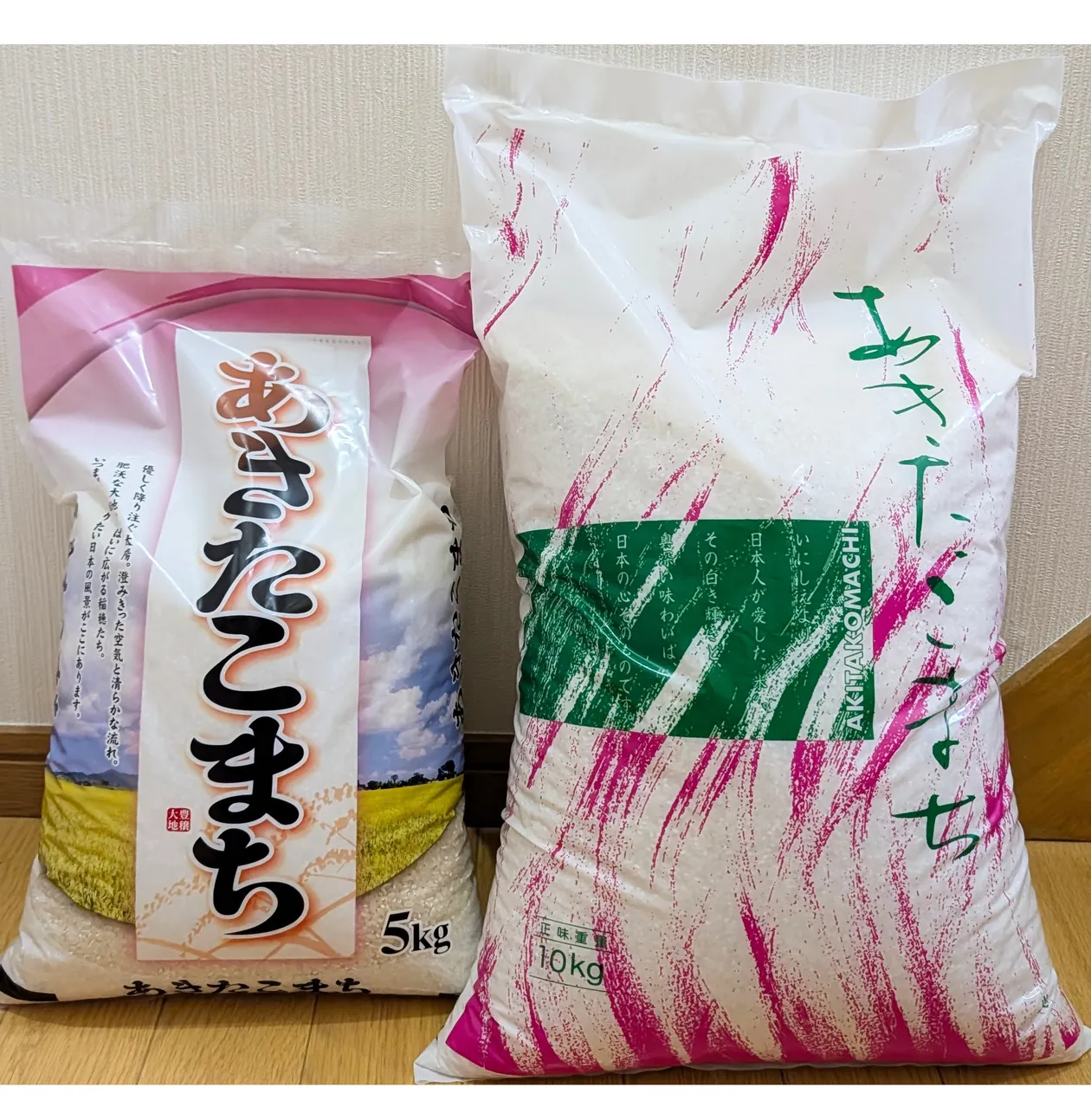 白い壁を背に立てて並べた、あきたこまちの5kg袋（左・ピンク基調のパッケージ）と10kg袋（右・白地にピンクの筆跡と緑の帯）。